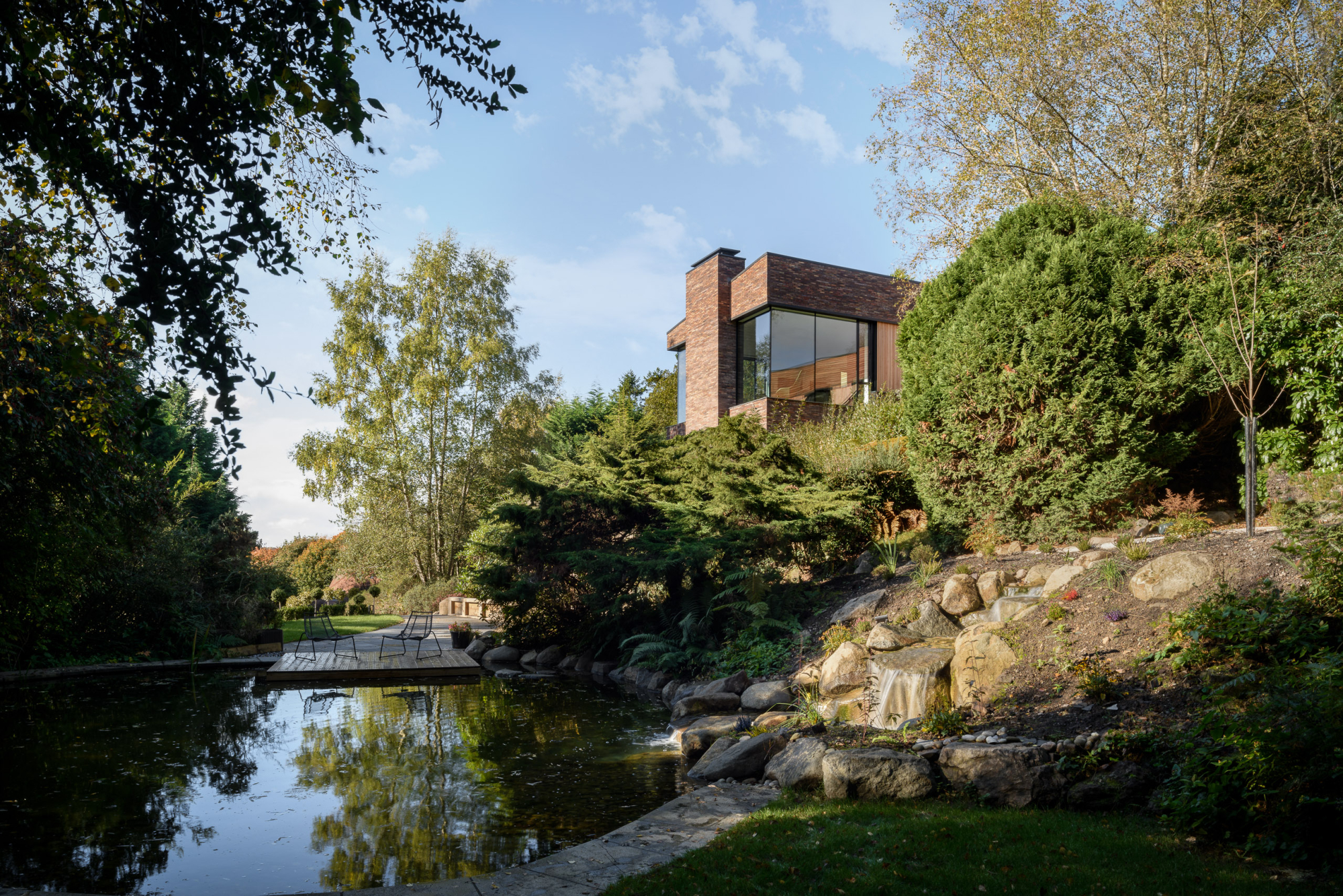 Modern house overlooking a pond with lush greenery and rocks, set in a peaceful natural landscape.